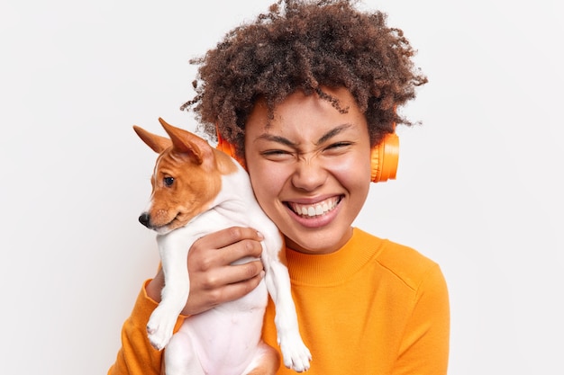 Concepto de amistad de animales de personas. Mujer de pelo rizado llena de alegría sonríe tiernamente sostiene a un pequeño cachorro de pedigrí cerca de la cara escucha música a través de auriculares inalámbricos aislados sobre una pared blanca