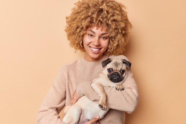 Foto gratuita concepto de amistad sonriente hermosa mujer joven lleva a su perro pug pasa tiempo libre con su mascota favorita usa un puente casual aislado sobre fondo marrón animal doméstico con su cariñoso dueño