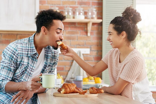 Concepto de amor y cuidado. Encantadora pareja se divierten juntos: mujer cariñosa alimenta a marido con croissant,