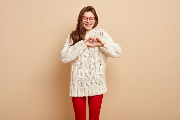 Concepto de amor. Feliz novia sonriente hace gesto de corazón con los dedos, expresa buenos sentimientos, vestida con un jersey blanco, aislado sobre una pared beige. Concepto de lenguaje de señas. Símbolo de la caridad