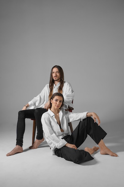 Concepto de amor, relaciones, moda y estilo. Imagen vertical aislada de pareja de moda en ropa similar posando descalzo. Hombre dominante relajándose en una silla con su esposa sentada en el piso