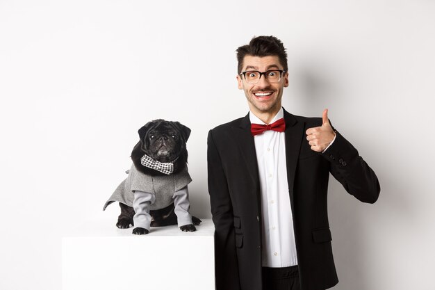 Concepto de animales, fiesta y celebración. Apuesto joven con traje y lindo pug negro en traje mirando a la cámara, propietario mostrando el pulgar hacia arriba en señal de aprobación y alabanza, fondo blanco.