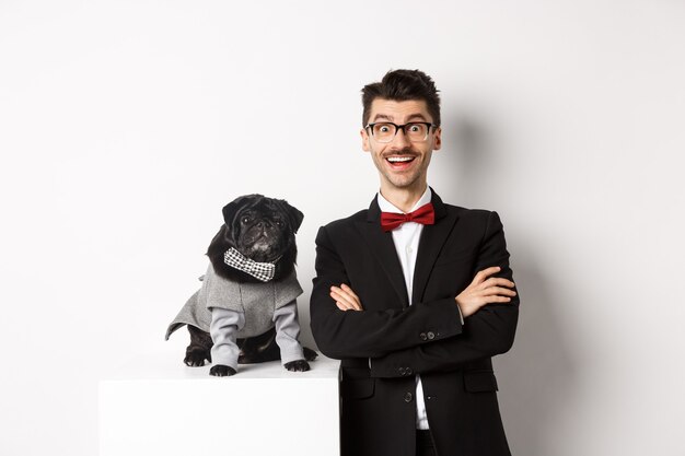 Concepto de animales, fiesta y celebración. Dueño de perro feliz en traje y cachorro en traje mirando emocionado a la cámara, divirtiéndose, de pie sobre fondo blanco.