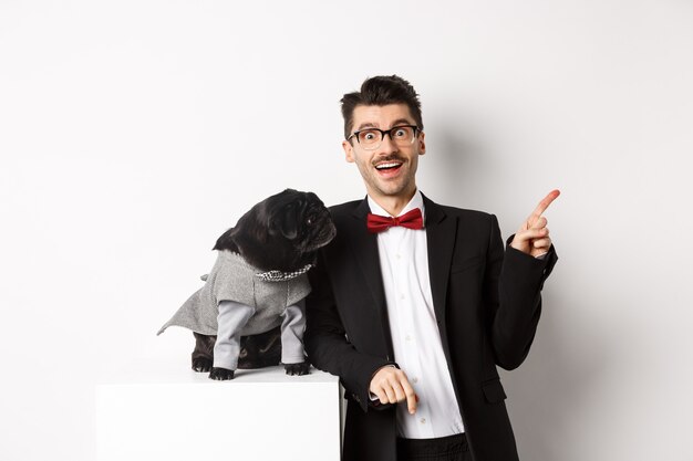 Concepto de animales, fiesta y celebración. Feliz y sorprendido joven y lindo pug en traje de pie sobre fondo blanco, dueño del perro apuntando a la derecha