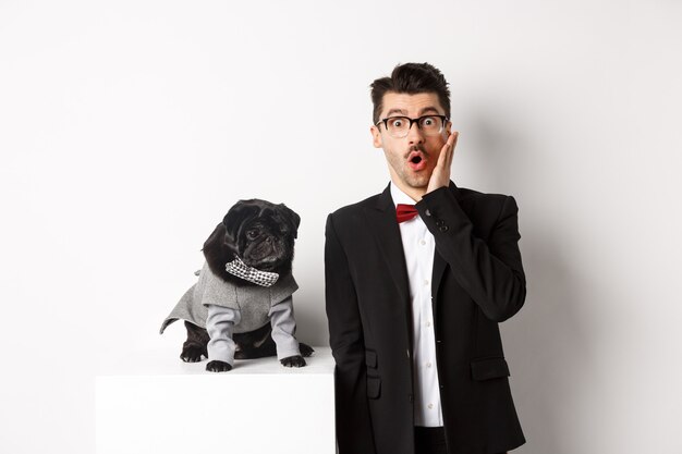 Concepto de animales, fiesta y celebración. Hombre guapo sorprendido en traje formal y lindo perro disfrazado, mirando a la cámara asombrado, de pie sobre fondo blanco.