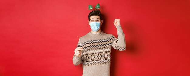 Concepto de año nuevo, covid-19 y distanciamiento social. Hombre feliz celebrando con máscara médica, sosteniendo bengalas y disfrutando de la fiesta, parado sobre fondo rojo.