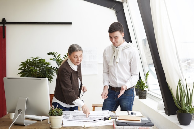 Concepto de arquitectura, ingeniería y diseño. Imagen del arquitecto hombre morena feliz sonriendo mientras su experimentada colega senior lo ayuda con su proyecto de construcción, enmendando dibujos