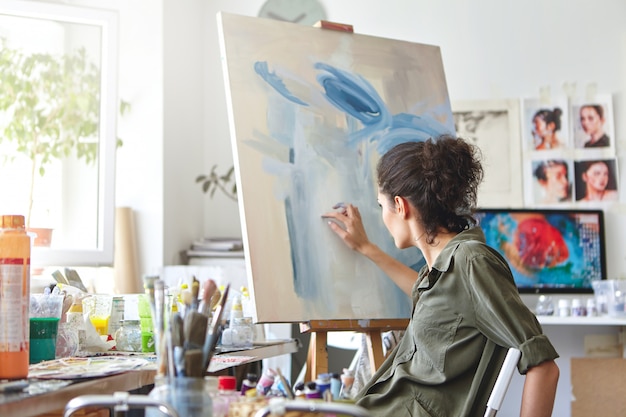 Concepto de arte, creatividad, hobby, trabajo y ocupación creativa. Vista posterior de la ocupada artista femenina sentada en una silla frente al caballete, pintando con los dedos, utilizando pintura al óleo o acrílica blanca y azul