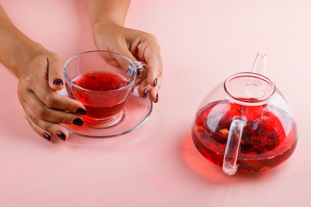 Concepto de bebida caliente con tetera en mesa rosa mujer sosteniendo la taza de té de cristal.