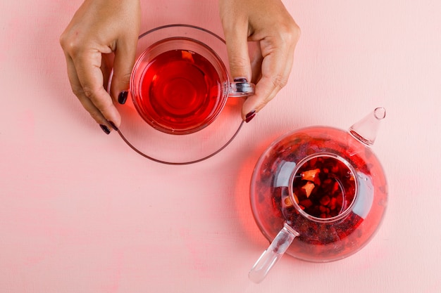 Concepto de bebida caliente con tetera en mesa rosa plana lay. mujer sosteniendo una taza de té de cristal.