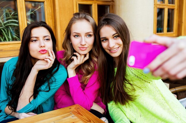 Concepto de bebidas, amistad, tecnología y personas: tres mujeres bonitas felices con tazas sentadas en la mesa y tomando selfie con smartphone en café. Colores brillantes y soleados.