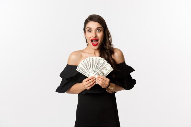 Concepto de belleza y compras. Mujer emocionada en vestido negro, mostrando el premio en dinero y mirando feliz a la cámara, de pie sobre fondo blanco.