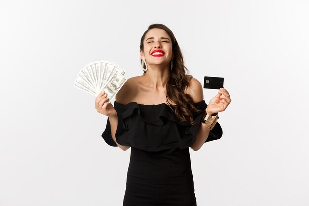 Concepto de belleza y compras. Mujer joven satisfecha con vestido elegante, mirando complacido, sosteniendo dinero y tarjeta de crédito, de pie sobre fondo blanco.