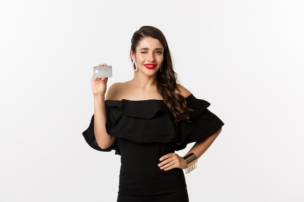 Concepto de belleza y compras. Mujer de moda en vestido negro, con maquillaje, guiñando un ojo a la cámara y mostrando la tarjeta de crédito, de pie sobre fondo blanco.