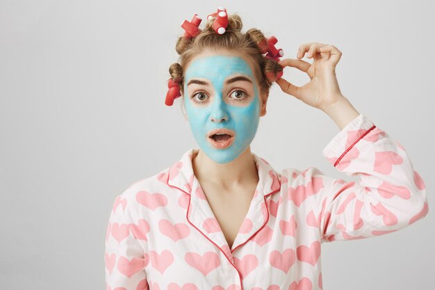 Concepto de belleza y cuidado de la piel. Bonita mujer en ropa de dormir y rizadores de pelo de despegue de máscara facial