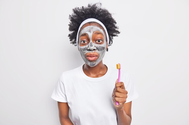 Concepto de belleza y cuidado de la piel. Mujer afroamericana sorprendida con cabello rizado aplica mascarilla de arcilla hidratante sostiene cepillo de dientes mira conmocionado a la cámara