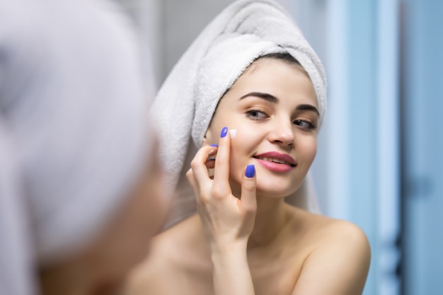 Concepto de belleza, cuidado de la piel y personas - mujer joven sonriente aplicando crema en la cara y mirando al espejo en el baño de casa