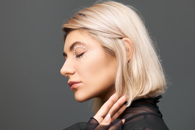 Concepto de belleza, estilo, moda, glamour y feminidad. Mujer joven con estilo de moda con peinado bob teñido, anillo en la nariz y cristales blancos alrededor de su ojo, posando aislado con los ojos cerrados