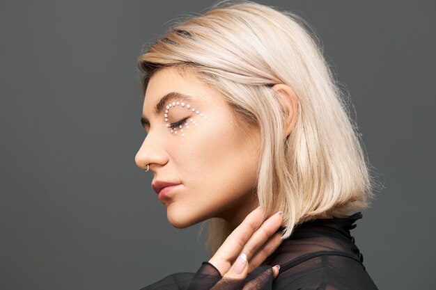 Concepto de belleza, estilo, moda, glamour y feminidad. Mujer joven con estilo de moda con peinado bob teñido, anillo en la nariz y cristales blancos alrededor de su ojo, posando aislado con los ojos cerrados