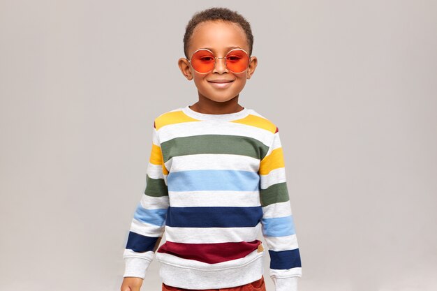 Concepto de belleza, estilo y moda. Imagen de niño africano de moda alegre posando aislado vistiendo elegante suéter a rayas y gafas de sol rosa redondas de moda, sonriendo felizmente