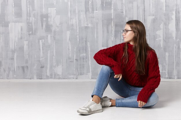 Concepto de belleza, estilo, moda, juventud, personas y estilo de vida. Bonita y encantadora adolescente con largo cabello suelto sentada en el piso, con gafas, suéter de cuello alto, jeans y zapatillas