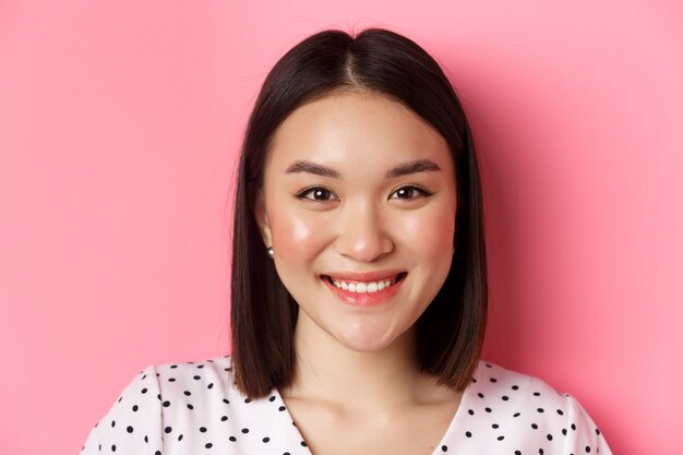 Concepto de belleza y estilo de vida. Foto de cabeza de hermosa mujer asiática sonriendo, mirando a cámara feliz y romántica, de pie contra el fondo rosa.