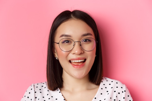 Concepto de belleza y estilo de vida. Primer plano de modelo de mujer asiática linda con gafas de moda, sonriendo feliz a la cámara, de pie sobre fondo rosa.