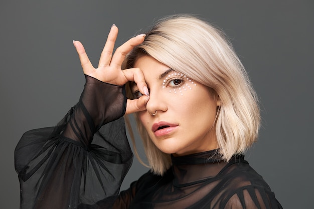 Concepto de belleza, glamour, lujo y moda. Foto de perfil de atractiva mujer joven fresca en elegante blusa negra transparente con bengalas posando aislado, conectando el pulgar y el dedo índice en señal de ok