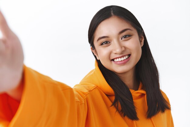 Concepto de belleza y moda de Asia Retrato de primer plano hermosa chica asiática con capucha naranja tomando selfie sosteniendo la cámara con la mano extendida y sonriendo mientras hace que la foto use nuevos filtros kawaii sonriendo