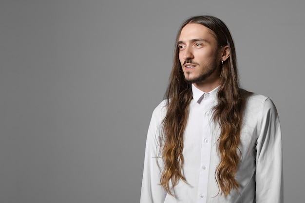 Concepto de belleza, moda, estilo y personas. Disparo aislado de extraordinario hombre joven de moda con pelo largo suelto, barba y pendiente posando en la pared gris, vistiendo elegante camisa blanca, sonriendo