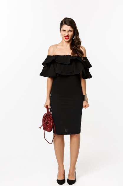 Concepto de belleza y moda. Longitud total de mujer enojada en vestido de fiesta negro y tacones altos, expresan desdén y muecas ante la cámara, enojado con la persona, fondo blanco.