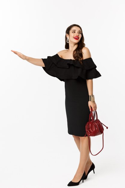 Concepto de belleza y moda. Longitud total de mujer glamour en vestido negro y tacones altos levantando la mano para detener un taxi, necesita un paseo, de pie sobre fondo blanco.