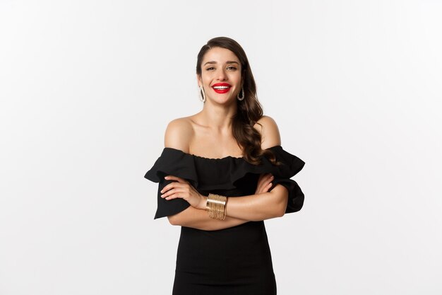 Concepto de belleza y moda. Modelo femenino atractivo en vestido de fiesta y lápiz labial rojo, sonriendo complacido, mirando feliz, de pie sobre fondo blanco.