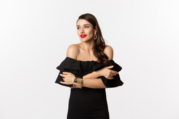 Concepto de belleza y moda. Mujer elegante y hermosa en vestido negro, maquillaje, abrazándose a sí misma y mirando a otro lado con mirada sensual, de pie sobre fondo blanco