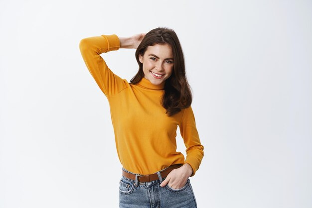 Concepto de belleza y moda Mujer joven con estilo en ropa de moda sosteniendo la mano detrás de la cabeza en una pose relajada y segura sonriendo a la cámara de fondo blanco