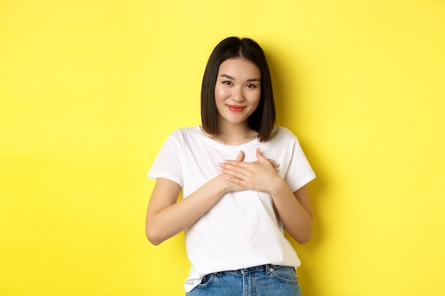 Concepto de belleza y moda. Sentida chica asiática cogidos de la mano en el corazón y sonriendo tocado, agradeciéndole, de pie sobre fondo amarillo