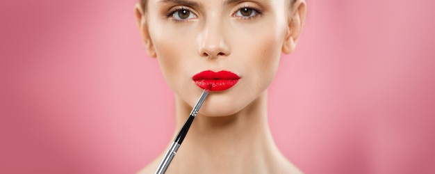 Concepto de belleza Mujer aplicando lápiz labial rojo con fondo de estudio rosa Hermosa chica hace maquillaje