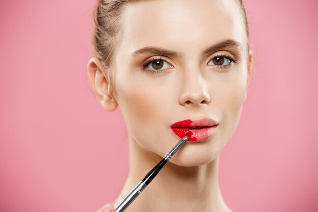 Concepto de belleza - mujer aplicar lápiz labial rojo con fondo de estudio de color rosa. Hermosa chica hace maquillaje.