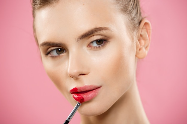 Concepto de belleza - mujer aplicar lápiz labial rojo con fondo de estudio de color rosa. Hermosa chica hace maquillaje.