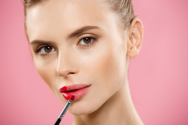 Concepto de belleza - mujer aplicar lápiz labial rojo con fondo de estudio de color rosa. Hermosa chica hace maquillaje.