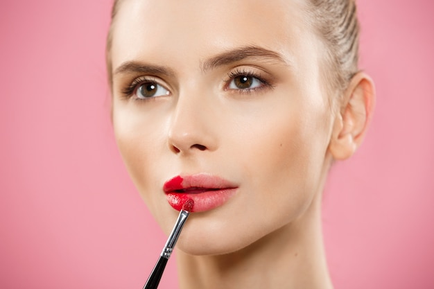 Concepto de belleza - mujer aplicar lápiz labial rojo con fondo de estudio de color rosa. Hermosa chica hace maquillaje.
