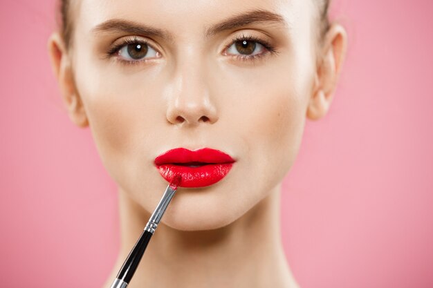 Concepto de belleza - mujer aplicar lápiz labial rojo con fondo de estudio de color rosa. Hermosa chica hace maquillaje.