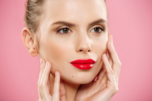 Concepto de belleza - mujer aplicar lápiz labial rojo con fondo de estudio de color rosa. Hermosa chica hace maquillaje.