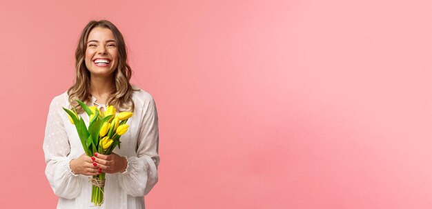 Concepto de belleza y primavera de vacaciones Retrato de feliz emocionada encantadora chica rubia recibe flores comprando tulipanes amarillos ella misma sonriendo y riendo alegremente de pie fondo rosa