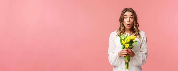 Concepto de belleza y primavera de vacaciones Retrato de niña rubia sorprendida y asombrada en vestido blanco sosteniendo tulipanes amarillos recibe flores siendo divertido y feliz de pie fondo rosa