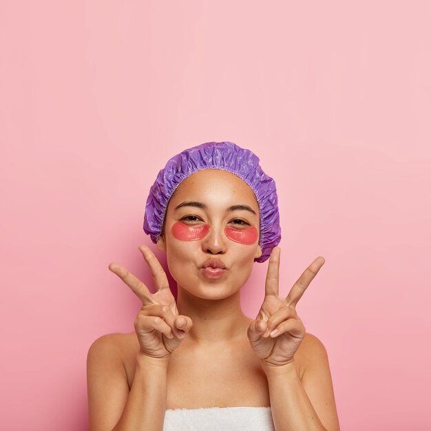 Concepto de belleza y rejuvenecimiento. Bonita mujer coreana hace un gesto de paz con la mano, mantiene los labios doblados, tiene parches debajo de los ojos en la cara, usa un sombrero de baño púrpura, disfruta de los procedimientos de spa después de tomar una ducha