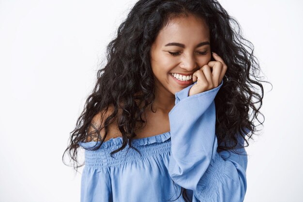 Concepto de belleza y ternura de bienestar Encantadora mujer afroamericana de pelo rizado con blusa azul riéndose tocando la mejilla y cerrando los ojos riéndose despreocupada de una broma graciosa de fondo blanco