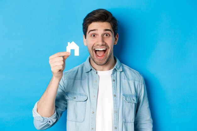 Concepto de bienes raíces. Feliz joven mirando emocionado, encontró apartamento, mostrando modelo de casa pequeña, de pie sobre fondo azul.