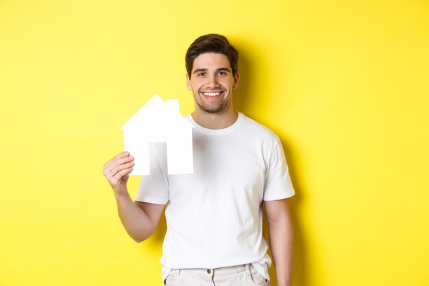 Concepto de bienes raíces. Hombre joven en camiseta blanca con modelo de casa de papel y sonriendo, buscando apartamento, de pie sobre fondo amarillo.