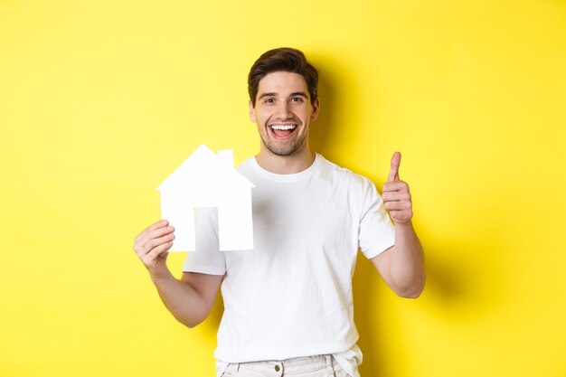 Concepto de bienes raíces. Hombre joven feliz que muestra el modelo de la casa de papel y los pulgares para arriba, recomendando el corredor, colocándose sobre fondo amarillo.